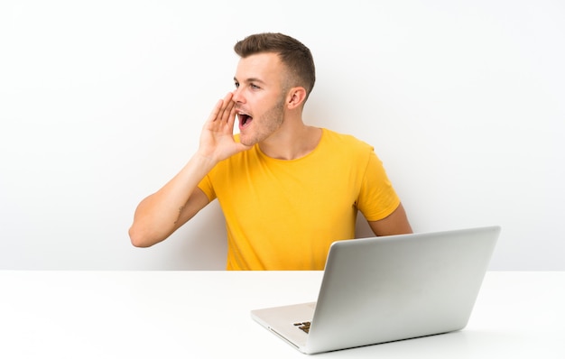 Junger blonder Mann in einer Tabelle mit einem Laptop, der mit dem breiten Mund schreit, öffnen sich