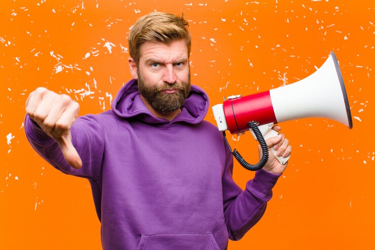 Foto junger blonder mann mit einem megaphon, das einen purpurroten hoodie gegen schädigende orange wand trägt