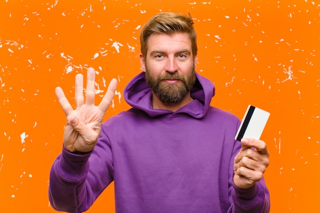 Junger blonder Mann mit einer Kreditkarte, die einen purpurroten Hoodie gegen schädigende orange Wand trägt