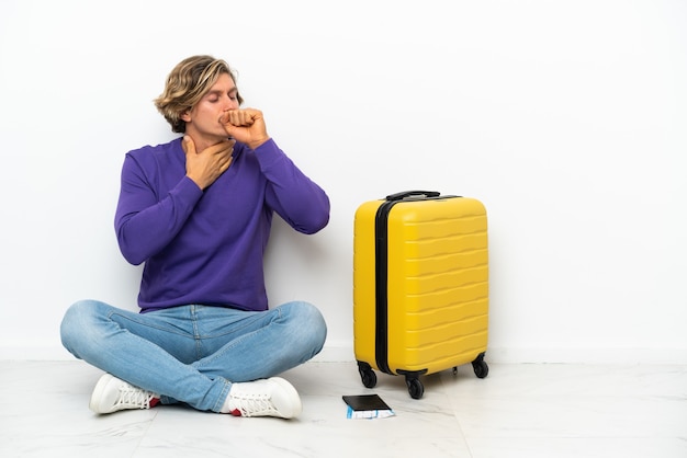 Junger blonder Mann mit Koffer, der auf dem Boden sitzt, leidet unter Husten und fühlt sich schlecht