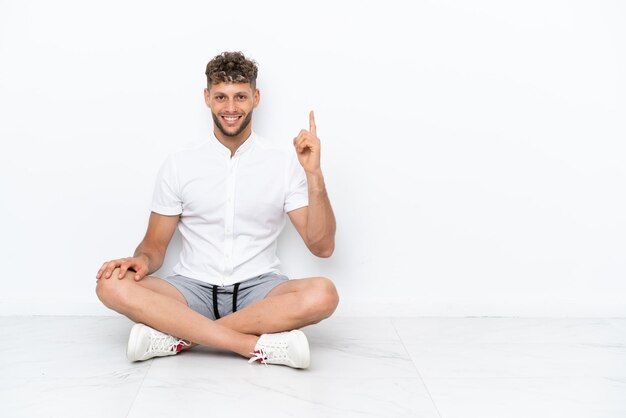Junger blonder Mann sitzt isoliert auf weißem Hintergrund auf dem Boden und zeigt und hebt einen Finger als Zeichen der Besten