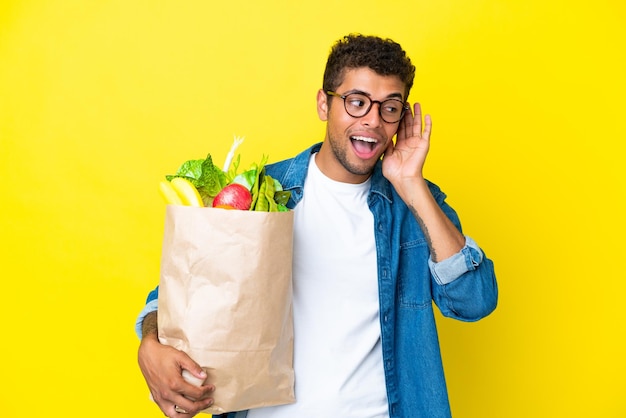 Junger Brasilianer, der eine Einkaufstasche isoliert auf gelbem Hintergrund hält und etwas hört, indem er die Hand auf das Ohr legt