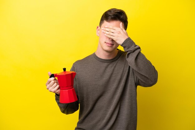 Junger brasilianer, der kaffeekanne isoliert auf gelbem hintergrund hält, der die augen mit den händen bedeckt, will nichts sehen