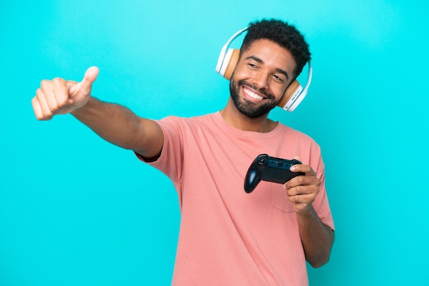 Junger Brasilianer, der mit einem Videospiel-Controller spielt, isoliert auf blauem Hintergrund, mit einer Daumen-nach-oben-Geste