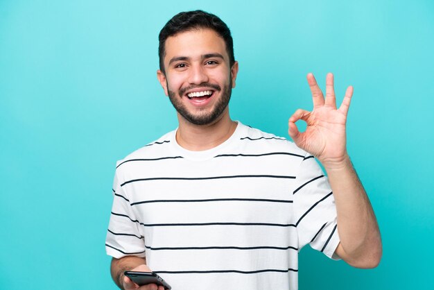 Junger Brasilianer isoliert auf blauem Hintergrund mit Handy und OK-Zeichen