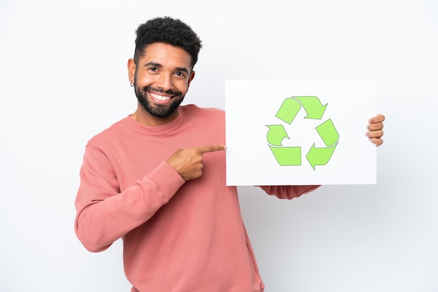 Junger Brasilianer isoliert auf weißem Hintergrund, der ein Schild mit Recycling-Symbol hält und darauf zeigt