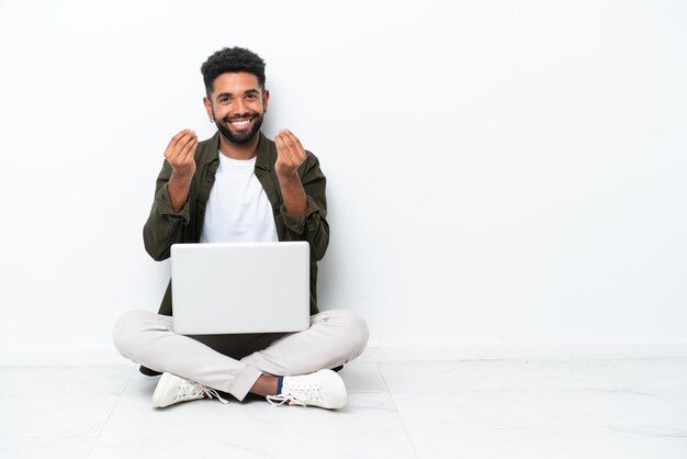 Junger Brasilianer mit einem Laptop, der auf dem Boden sitzt, isoliert auf einer weißen Geste, Geld zu verdienen