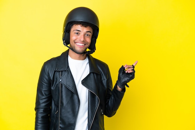 Junger Brasilianer mit einem Motorradhelm isoliert auf gelbem Hintergrund, der zur Seite zeigt, um ein Produkt zu präsentieren