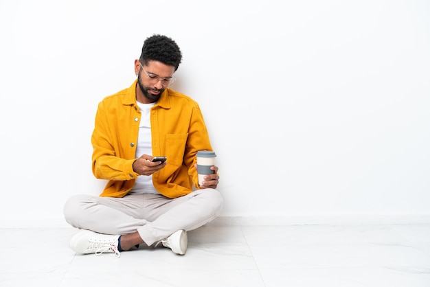 Junger Brasilianer sitzt isoliert auf weißem Hintergrund auf dem Boden und hält Kaffee zum Mitnehmen und ein Handy