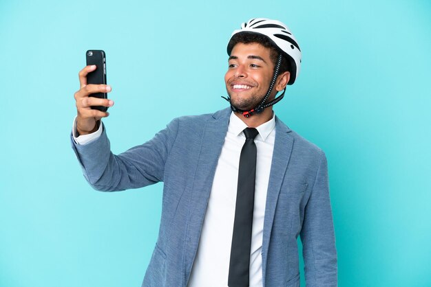 Junger brasilianischer Geschäftsmann mit Fahrradhelm isoliert auf blauem Hintergrund, der ein Selfie macht