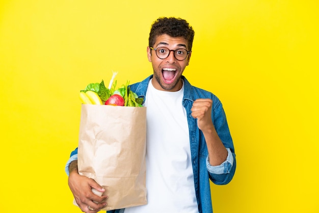 Junger brasilianischer Mann, der eine Einkaufstüte isoliert auf gelbem Hintergrund hält und einen Sieg in Siegerposition feiert