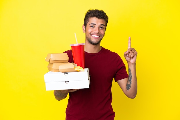 Junger brasilianischer Mann, der Pizza und Burger hält, isolierter Hintergrund, der einen Finger im Zeichen des Besten zeigt und hebt