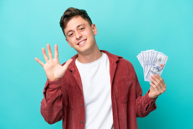 Junger brasilianischer mann, der viel geld nimmt, isoliert auf blauem hintergrund, der fünf mit den fingern zählt