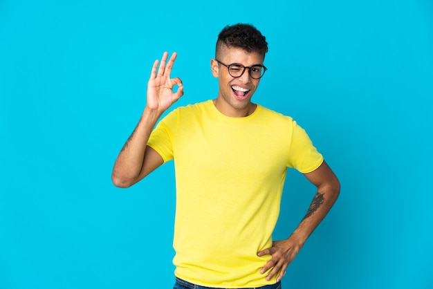 Junger brasilianischer Mann lokalisiert auf blauem Hintergrund, der ok Zeichen mit den Fingern zeigt