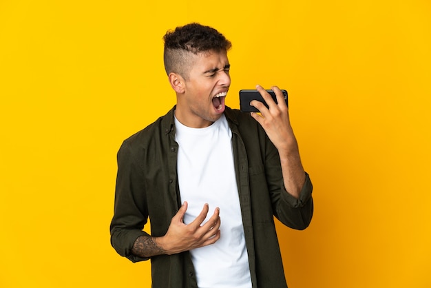 Junger brasilianischer Mann über isolierter Wand mit Handy und Gesang
