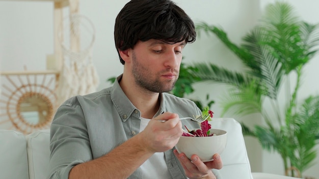 Junger brünetter Mann, der frischen Gemüsesalat isst, während auf der Couch zu Hause sitzt.