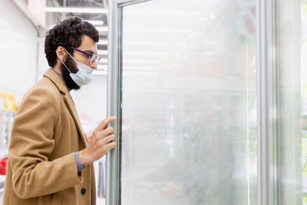 Junger brünetter mann in einer medizinischen maske im supermarkt in der abteilung mit tiefkühlkost. coronavirus pandemie. platz für text.