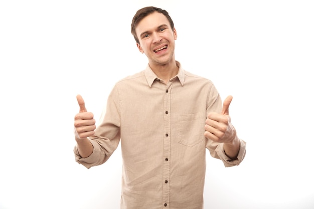 Junger brünetter Mann zeigt Hand mit erhobenem Daumen und lächelt isoliert auf weißem Hintergrund in die Kamera