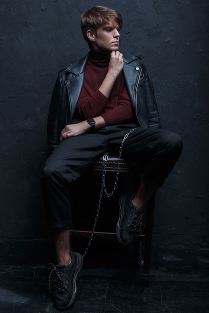 Junger charmanter Mann mit einer Frisur in einer Retro-Jacke im roten Golf in stilvollen Jeans in schwarzen Turnschuhen mit einer silbernen Metallkette sitzt und posiert auf einem Holzstuhl in einem dunklen Studio. Stilvoller Typ