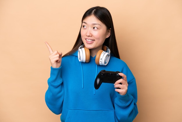 Junger Chinese, der mit einem Videospiel-Controller spielt, isoliert auf beigem Hintergrund, der auf eine großartige Idee zeigt
