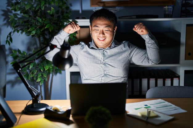Junger chinesischer Mann, der nachts mit einem Computer-Laptop arbeitet und Armmuskeln zeigt, lächelt stolz auf sein Fitnesskonzept