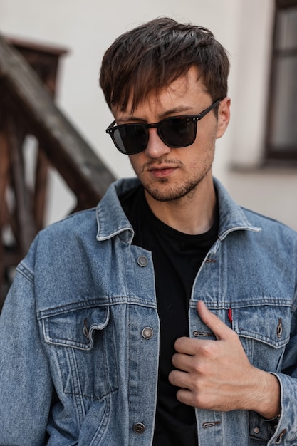 Junger cooler Mann richtet gerade modische blaue Jeansjacke gerade aus. Attraktiver urbaner Kerl-Hipster in der trendigen Sonnenbrille in der stilvollen Frühlingskleidung, die in der Stadt nahe einem Vintagen Gebäude aufwirft. Streetstyle. Herrenbekleidung