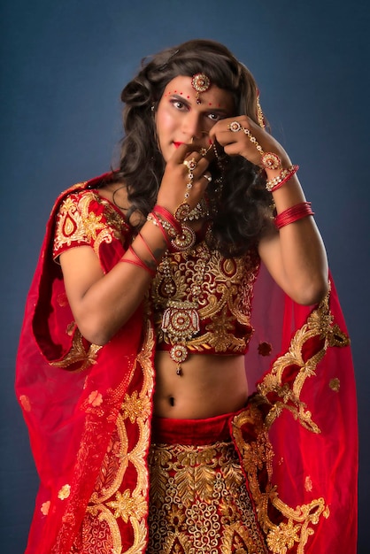 Junger Crossdresser-Mann, der indische Hochzeitskleidung einer Braut mit Braut-Make-up trägt und auf grauem Hintergrund posiert und modisch und glamourös aussieht