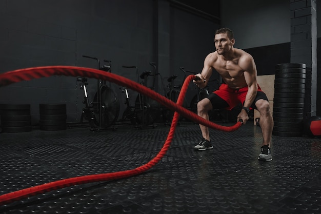 Junger Crossfit-Athlet, der Kampfseilübung an der Turnhalle tut. Manntrainingstraining mit Seil. Das Konzept der Sportmotivation. Speicherplatz kopieren.