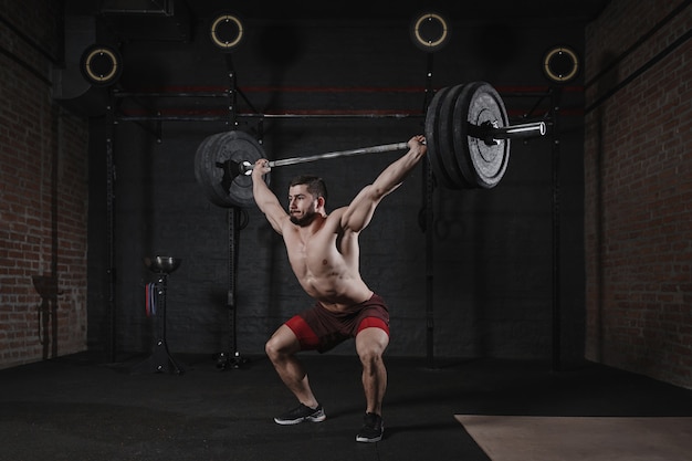 Junger Crossfit-Athlet, der schwere Langhantel über Kopf an der Turnhalle hebt. Hübscher Mann, der funktionelles Training tut. Powerlifting üben.