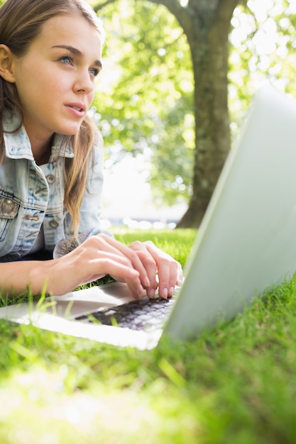 Junger denkender Student, der auf dem Gras unter Verwendung ihres Laptops liegt