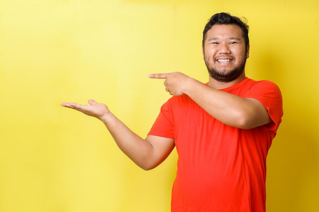 Junger dicker asiatischer Mann in Freizeitkleidung mit einem breiten Lächeln im Gesicht, das mit Hand und Finger t zeigt