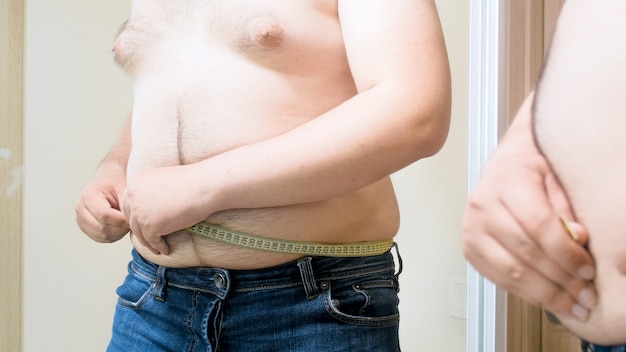 Junger dicker Mann, der großen Bauch mit Maßband misst. Konzept von männlichem Übergewicht, Gewichtsverlust und Diät.