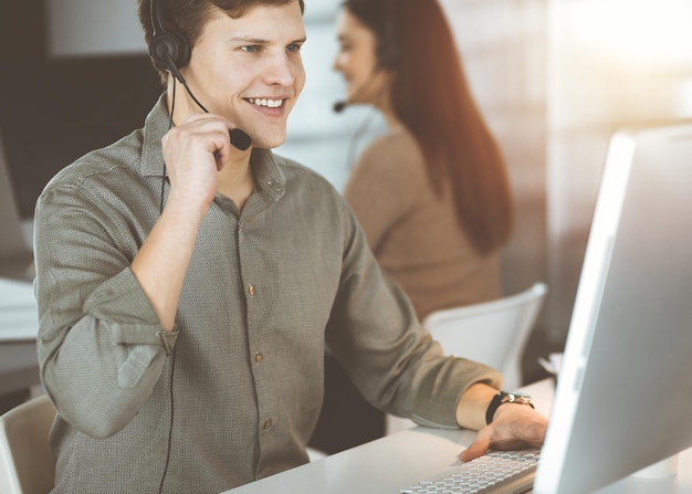 Junger dunkelhaariger Typ mit Kopfhörern spricht mit einem Kunden, während er in einem sonnigen Büro sitzt. Call-Center-Betreiber bei der Arbeit.