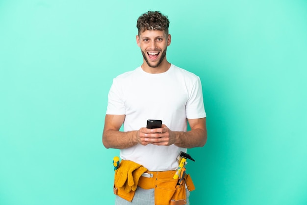 Junger Elektriker blonder Mann isoliert auf grünem Hintergrund überrascht und sendet eine Nachricht