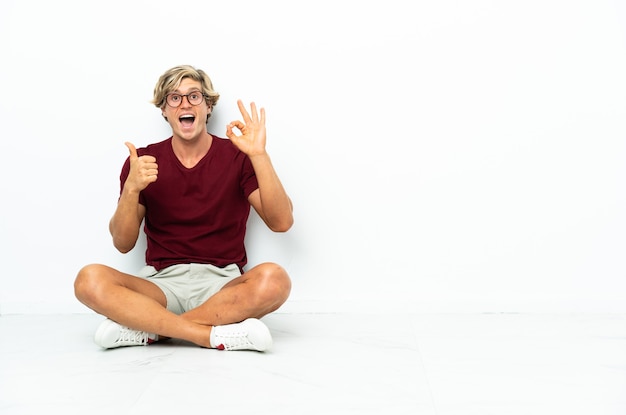 Junger englischer Mann, der auf dem Boden sitzt und ok Zeichen und Daumen hoch Geste zeigt