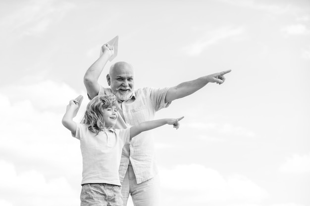 Junger Enkel und alter Großvater spielen mit Spielzeugpapierflugzeug vor Sommerhimmelhintergrund