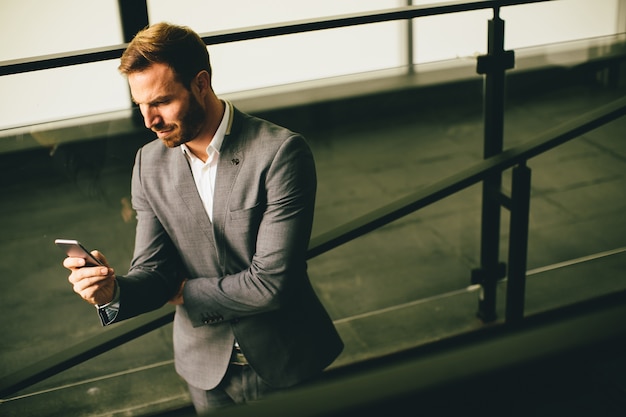 Junger erfolgreicher Geschäftsmann, der grauen Anzug trägt und seinen Smartphone bei nahe modernes stehen hält