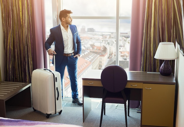 Junger erfolgreicher Geschäftsmann mit Gepäck, das Fenster im Hotelzimmer betrachtet.
