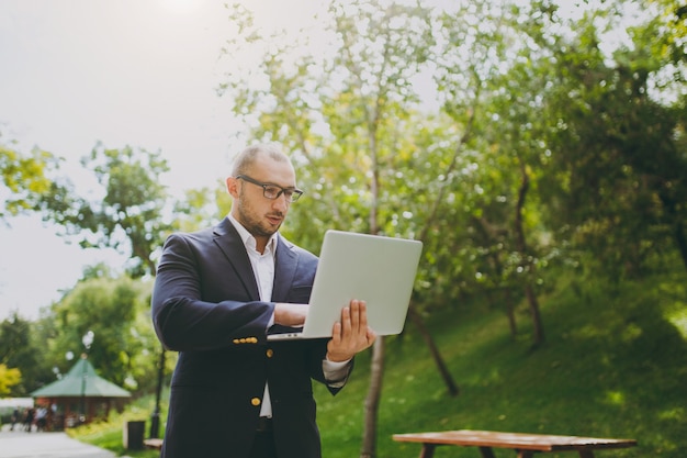 Junger erfolgreicher intelligenter Geschäftsmann im weißen Hemd, im klassischen Anzug, in der Brille. Mann steht und arbeitet an Laptop-PC im Stadtpark im Freien auf Naturhintergrund. Mobiles Büro, Geschäftskonzept.