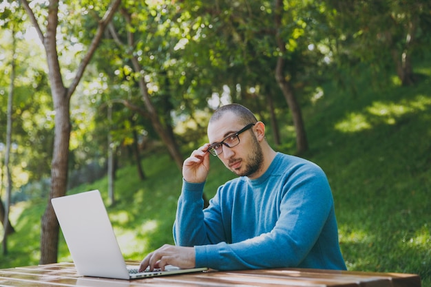 Junger erfolgreicher intelligenter Geschäftsmann oder Student in lässiger blauer Hemdbrille, der am Tisch mit Handy im Stadtpark sitzt und Laptop verwendet, der draußen auf grüner Natur arbeitet. Mobile Office-Konzept.