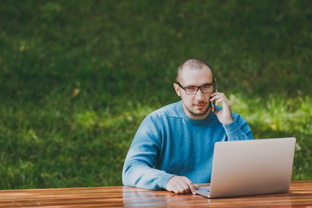 Junger erfolgreicher lächelnder intelligenter Manngeschäftsmann oder -student in lässigem blauem Hemd, Brille sitzt am Tisch, telefoniert im Stadtpark mit Laptop, arbeitet im Freien. Mobile Office-Konzept.