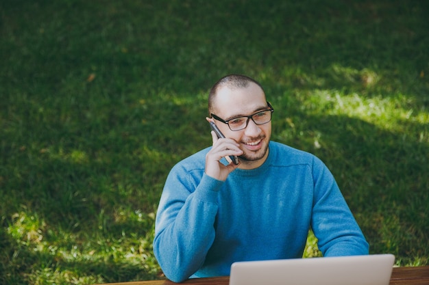 Junger erfolgreicher lächelnder intelligenter Manngeschäftsmann oder -student in lässigem blauem Hemd, Brille sitzt am Tisch, telefoniert im Stadtpark mit Laptop, arbeitet im Freien. Mobile Office-Konzept.