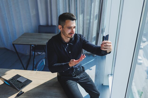 Junger erfolgreicher männlicher Arbeiter mit Brille und schwarzem Hemd, der auf dem Schreibtisch sitzt und über einen Laptop in einem modernen Büro an einem Projekt arbeitet und auf den Bildschirm blickt