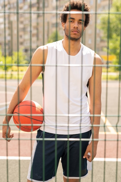 Junger ernsthafter aktiver Spieler mit Ball, der Sie durch Zaun des Basketballplatzes oder des Spielplatzes nach Spiel betrachtet