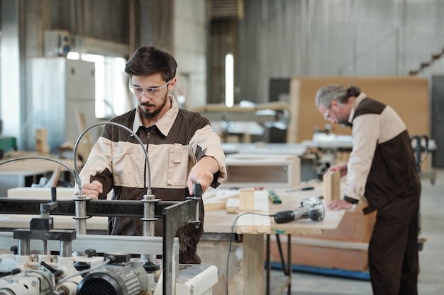 Junger ernsthafter Arbeiter der Möbelfabrik, der Sie während der Arbeit an der Werkbank an der Wand des Werkstattinnenraums ansieht