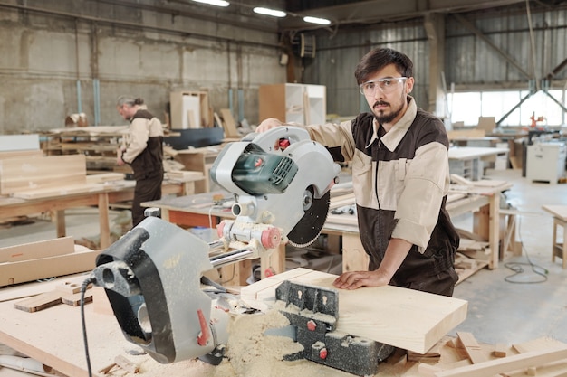 Junger ernsthafter Arbeiter oder Ingenieur der Möbelfabrik in Uniform und Brille, der in einer großen Werkstatt vor der Kamera steht