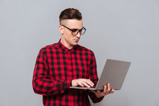 Junger ernsthafter Hipster in Brille und rotem Hemd, der Laptop in den Händen hält und etwas schreibt. Isolierter grauer Hintergrund