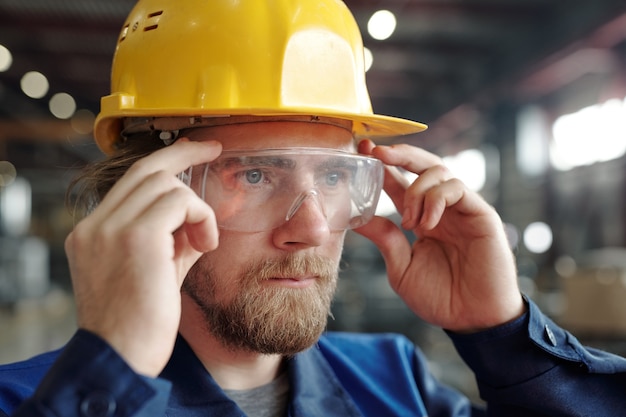 Junger ernsthafter männlicher Ingenieur oder Fabrikarbeiter in Schutzhelm und Brille, der in Werkstattumgebung vor der Kamera steht