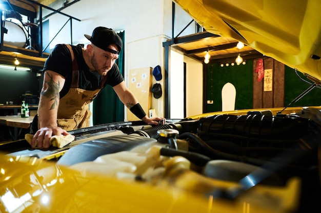 Junger ernsthafter Mann in Schürze und Baseballkappe, der sich über die offene Motorhaube des Autos beugt