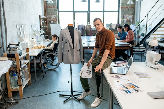 Junger ernsthafter Modedesigner oder Schneider mit Beinprothese, der von einer Schaufensterpuppe gegen beschäftigte weibliche Kollegen in einer großen Werkstatt auf dem Tisch sitzt